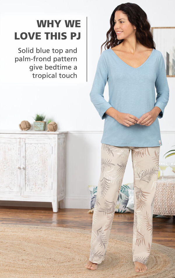 Model by bed wearing Margaritaville Pajamas - a solid blue 3/4 sleeve top with full-length pants in a cream palm-frond pattern that gives bedtime a tropical touch image number 2