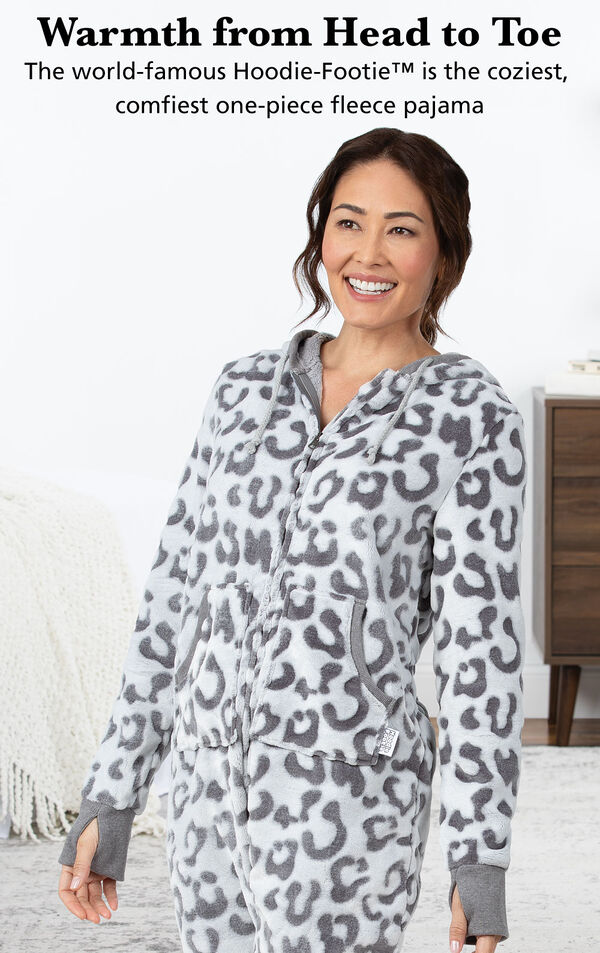 Model wearing Hoodie-Footie - Snow Leopard by bed with the following copy: Warmth from Head to Toe, The world famous Hoodie-Footie is the coziest, comfiest one-piece fleece pajama