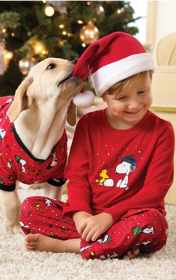 Boy sitting cross legged by Christmas Tree wearing Red Snoopy and Woodstock Toddler Pajamas and a Santa Hat, with his dog whom is wearing matching Snoopy Pajamas
