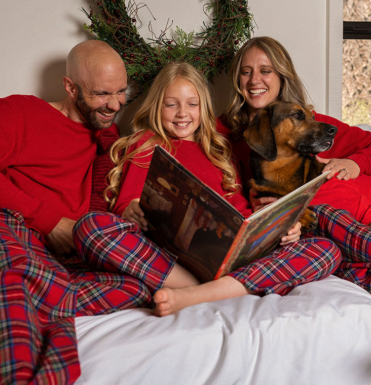 A family sitting in bed with their dog reading a book and wearing Stewart Plaid Flannel Matching Family Pajamas